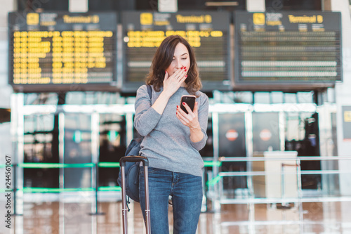 Image of excited, shocked young beautiful woman, standing in airport with smartphone in her hands - missed or cancelled flight concept - flight information board in background