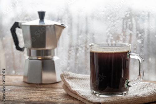 A cup of coffee on the background of geyser coffee machine and window with raindrops. Closeup, selective focus, wooden surface