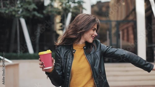 Happy young woman walking near the business center and dancing with a cup of coffee in hand. Beautiful girl looking into the camera. Good mood, funny, slow motion photo