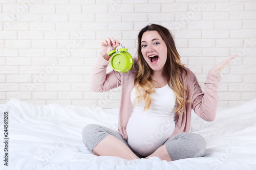 Beautiful pregnant woman with alarm clock sitting on bed