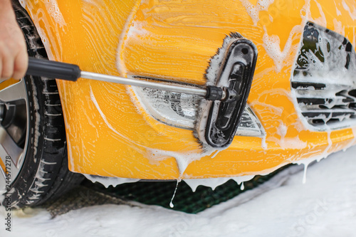 Front bumper and lights of yellow car washed in self serve carwash, brush moving in white shampoo.