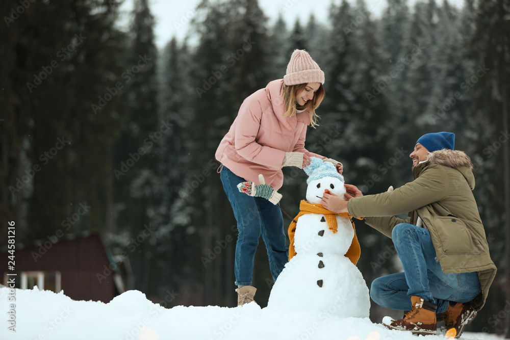 Fototapeta premium Couple making snowman near forest. Winter vacation