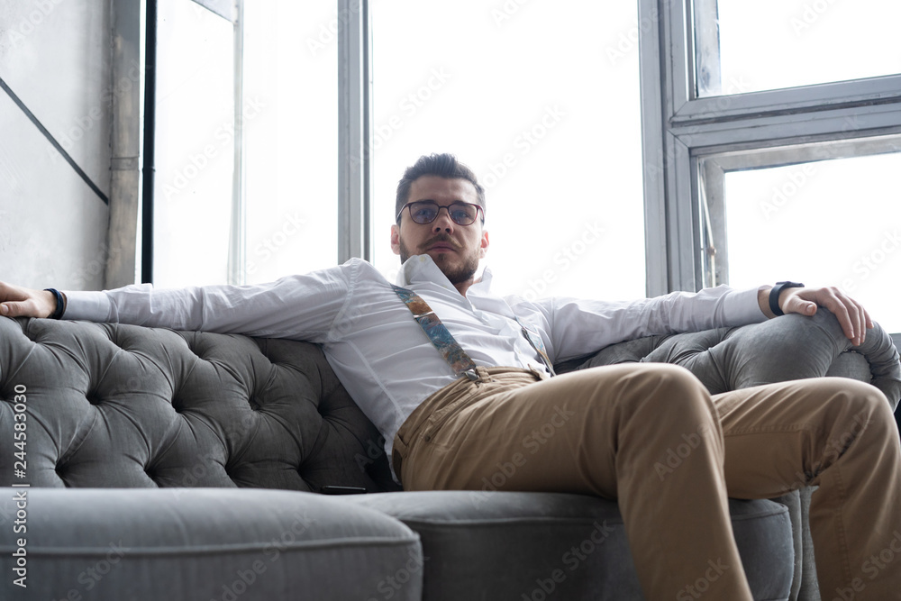 Enjoying free time. Thoughtful young man in full suit looking away while sitting on the sofa at home.