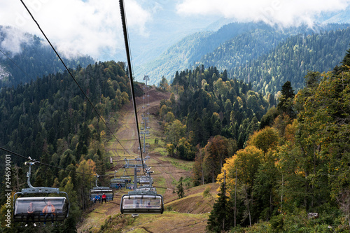 Cableway in the foreground. Mountains covered with forests. Yellow trees. Early autumn, sunny day. In the background low clouds, partially covering the mountains.