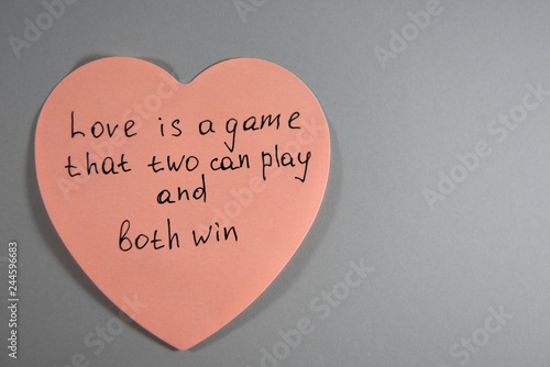 Pink sticker in the form of heart on a gray background with an inscription: Love is a game that two can play and both win