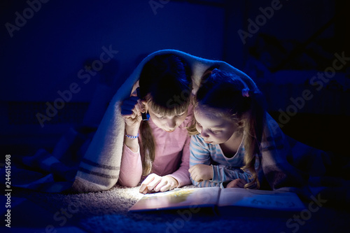 two sisters reading a book with a flashlight in a dark room at night
