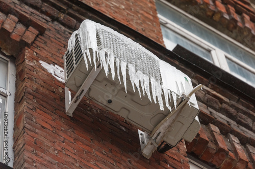 Air conditioning covered with ice and icicles on brick wall/danger for passers, сold winter, poor thermal insulation, ice stalactite, formation of icicles, frost and winter weather concept.