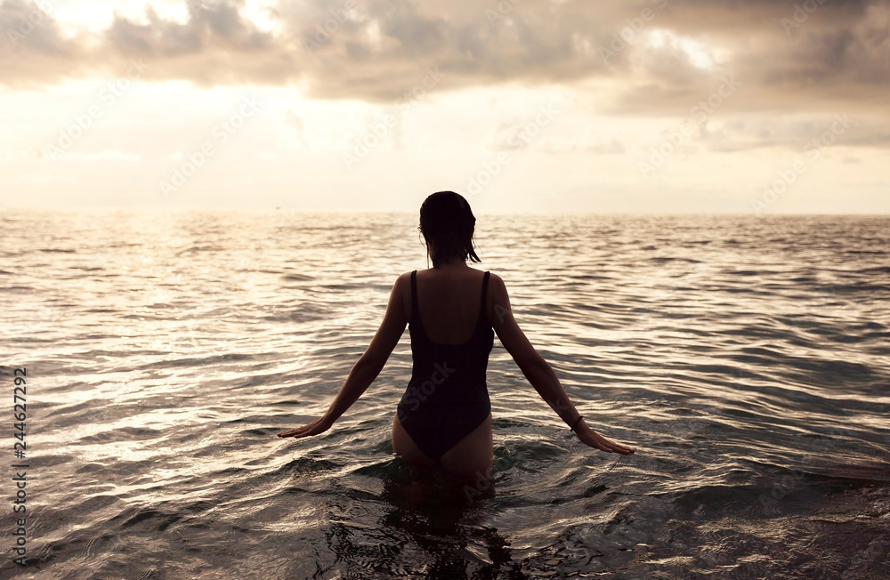 Young girl in warm water at sunset. Gorgeous colors in the sky and the sea. Perfect shapes reflected in water