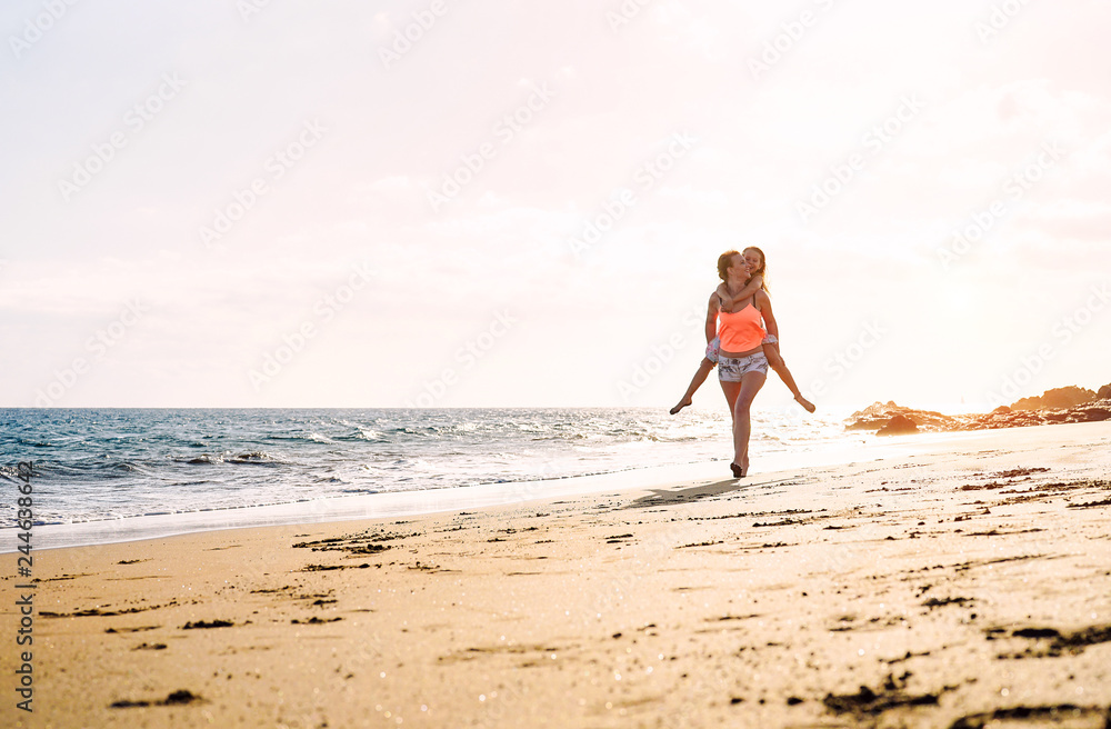 Happy loving family mother and daughter running and having fun on the beach at sunset - Mom piggyback her kid next to ocean during holidays - Parent, vacation, family lifestyle concept Happy loving family mother and daughter running and having fun on the beach at sunset - Mom piggyback her kid next to ocean during holidays - Parent, vacation, family lifestyle concept