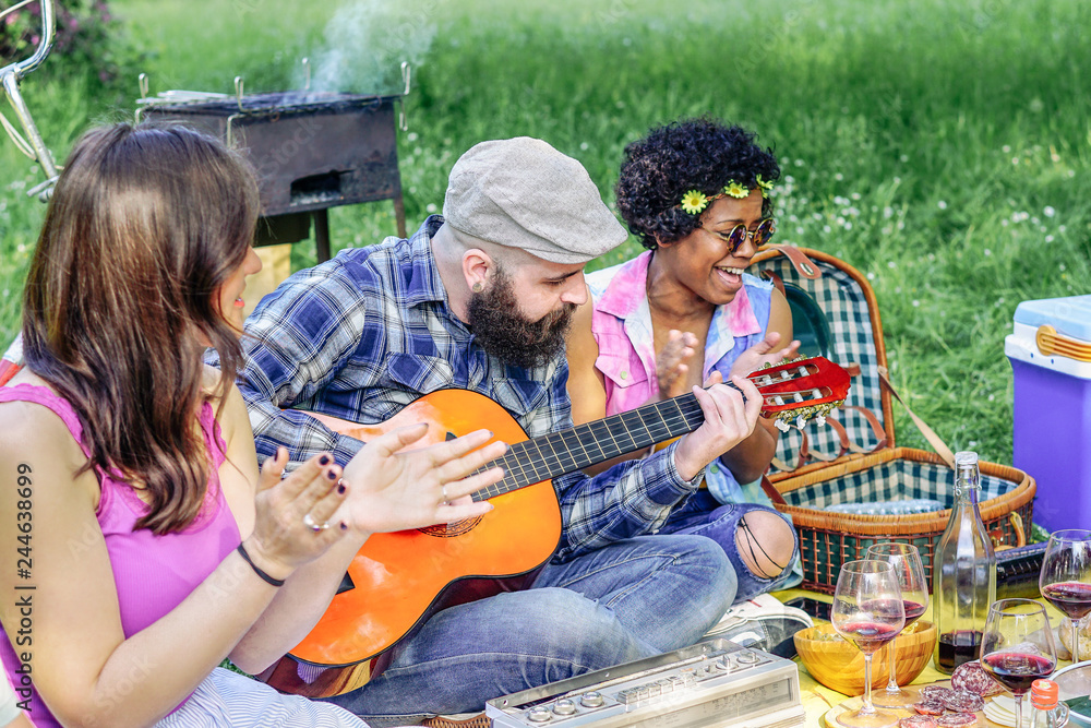 Happy friends playing guitar at picnic lunch in a park outdoor - Young people having fun and laughing together at barbecue in garden - Friendship, bbq and youth lifestyle concept