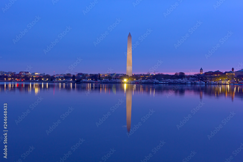 US capital panorama along Tidal Basin reservoir during cherry blossom at dawn. Easy recognizable Washington Monument with reflection in water stands tall on horizon.