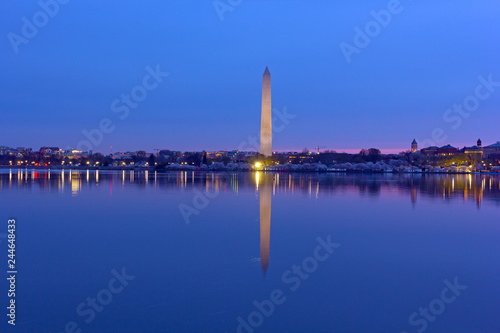 US capital panorama along Tidal Basin reservoir during cherry blossom at dawn. Easy recognizable Washington Monument with reflection in water stands tall on horizon.
