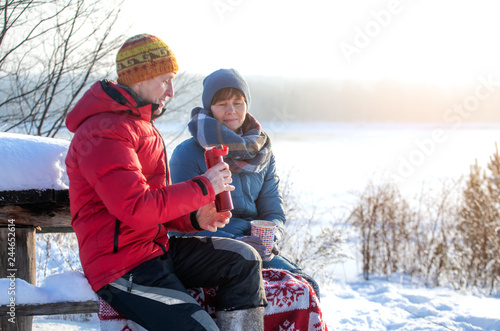 A man and a woman are drinking tea, coffee, drinks, sitting outdoors with a river view. Frosty sunny winter day