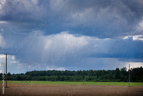 Rural landscape with rain storm over the meadow next to the forest. Straw bales standing in green  fresh grass during the monsoon and thunder storm. Dramatic sky  sunlight coming through dark clouds