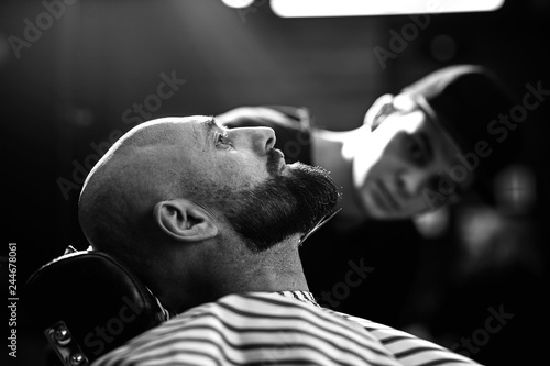 Black and white photo. Fashion barber dressed in a black clothes tidies up men's beard  and scissors it in the barbershop