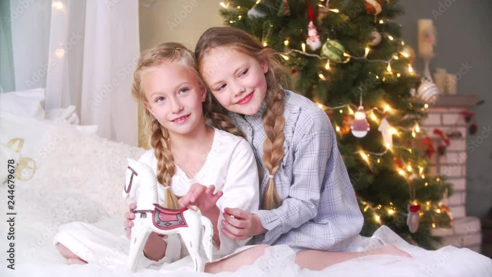 Two cute sisters with long braids and in nightwear are posing together with toy horse on big bed in the Christmas morning Two cute sisters with long braids and in nightwear are posing together with toy horse on big bed in the Christmas morning