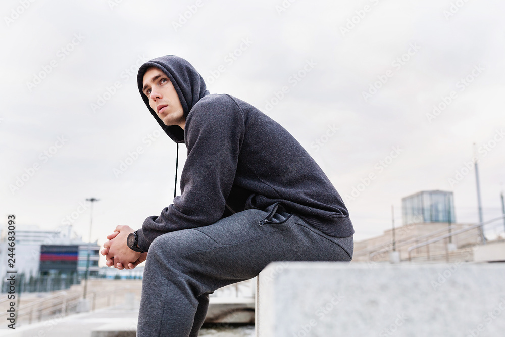 Tired young slim guy fitness blogger rests after a morning training in urban space. Concept of self-organization and building a slim body. Place for advertising content Tired young slim guy fitness blogger rests after a morning training in urban space. Concept of self-organization and building a slim body. Place for advertising content