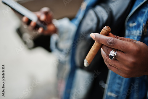 Close up hands of african american man in jeans jacket,  smoking cigar with mobile phone at hand. © AS Photo Family