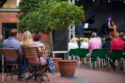 Married couple is sitting in restaurant, drinking wine, eating fancy snacks and listening to jazz concert in concert venue in city square. Musicians, stage, crowd and concert in the background.