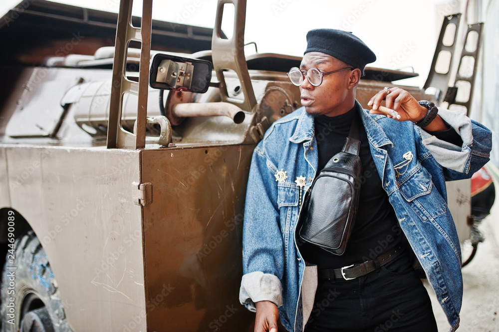 African american man in jeans jacket, beret and eyeglasses, smoking cigar and posed against btr military armored vehicle.