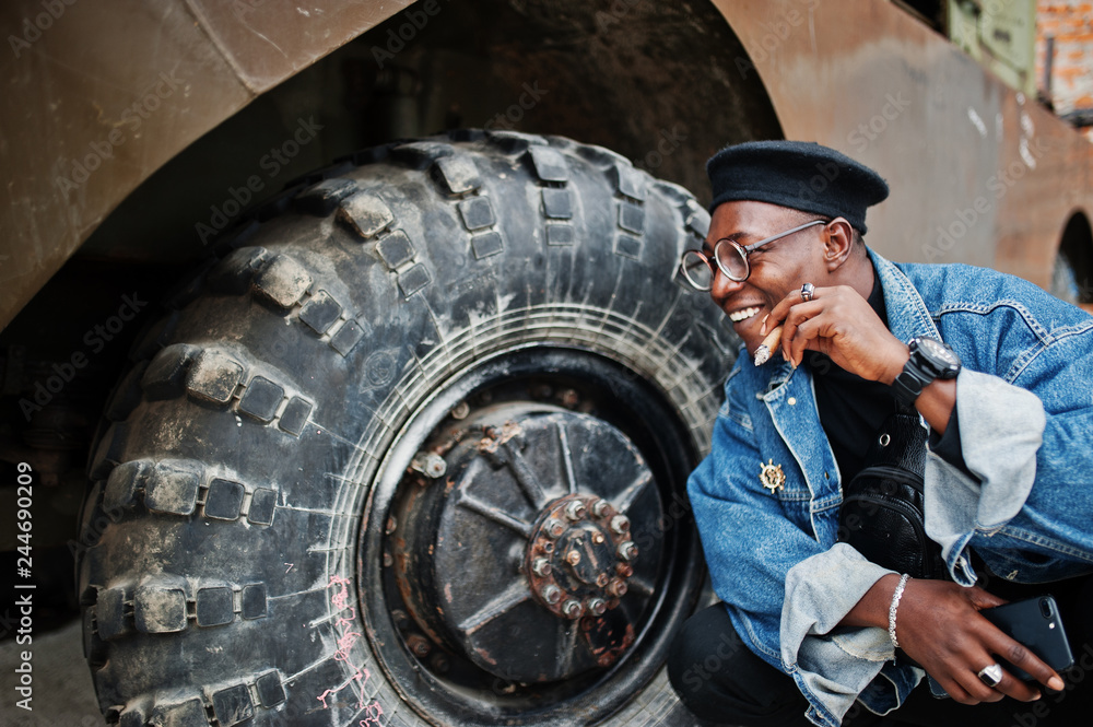 African american man in jeans jacket, beret and eyeglasses, smoking cigar and posed against wheel of btr military armored vehicle. African american man in jeans jacket, beret and eyeglasses, smoking cigar and posed against wheel of btr military armored vehicle.