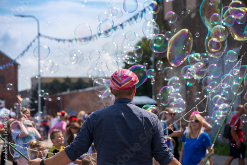 A freelance clown blowing hundreds of tiny, small and big bubbles at outdoor festival in city center. Concept of entertainment, birthdays. Kids having fun. Shower of bubbles flying in the happy crowd