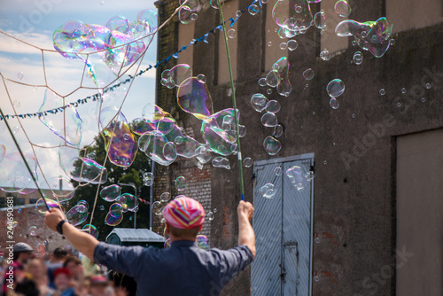 A freelance clown blowing hundreds of tiny, small and big bubbles at outdoor festival in city center. Concept of entertainment, birthdays. Kids having fun. Shower of bubbles flying in the happy crowd