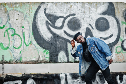 African american man in jeans jacket, beret and eyeglasses against graffiti wall with skull. © AS Photo Family
