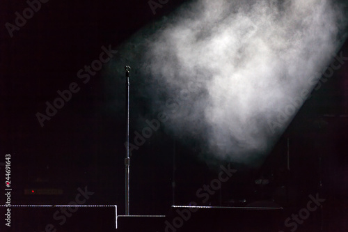 Microphone holder standing on a black dark scene, smoke illuminated by white spotlight at the side, below are stage monitors. Microphone stand on stage.