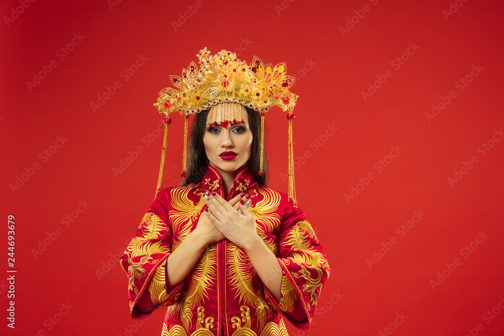 Fototapeta premium Chinese traditional graceful woman at studio over red background. Beautiful girl wearing national costume. Chinese New Year, elegance, grace, performer, performance, dance, actress, dress concept