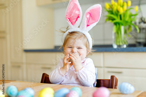 Cute little toddler girl wearing Easter bunny ears playing with colored pastel eggs. Happy baby child unpacking gifts. Adorable kid in pink clothes enjoying holiday