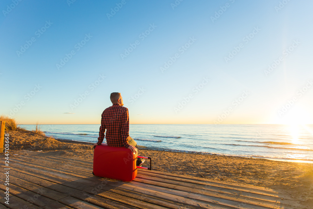 Fototapeta premium Holiday, travel and summer vacation concept -a man sitting on red suitcase and watching the sunset