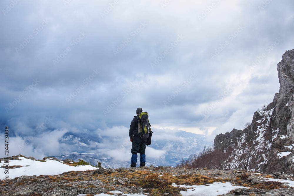 Fototapeta premium Hiker standing on a mountain top and looking into dynamic sky. Foggy mountain, Colorful winter landscape