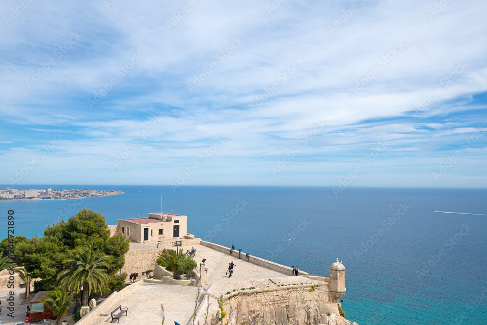 Panoramic view of city and Postiguet beach from Santa Barbara Castle in Alicante, Spain. Big palm trees in fortress on beautiful sea or ocean landscape in background, blue sky and tropical weather  