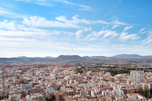 Panoramic view of city from Santa Barbara Castle in Alicante, Spain. Block apartment buildings, parks, roads, houses, palm trees. Beautiful mountain landscape in background, blue sky   © lainen