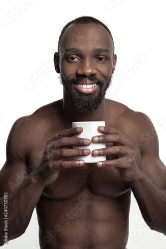African man with white cup of tea or coffee, isolated on white studio background. Close up portrait in minimalism style of a young naked happy afro man
