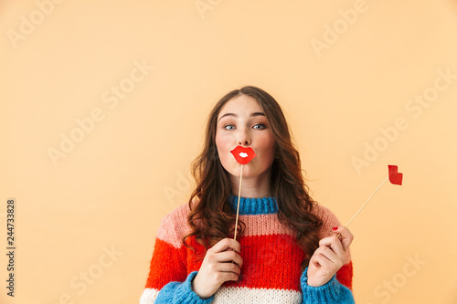Image of european woman 20s with long hair smiling and holding paper lips, standing isolated over beige background