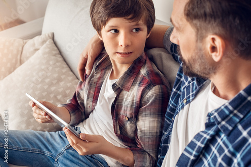 Father and little son at home sitting on sofa dad hugging boy with digital tablet looking at each other serious close-up
