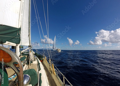 Across the teak deck of a traditional sailboat with full white sails, green canvas, and bronze and stainless fittings underway on wavy indigo seas traveling towards another sailing vessel passing by.