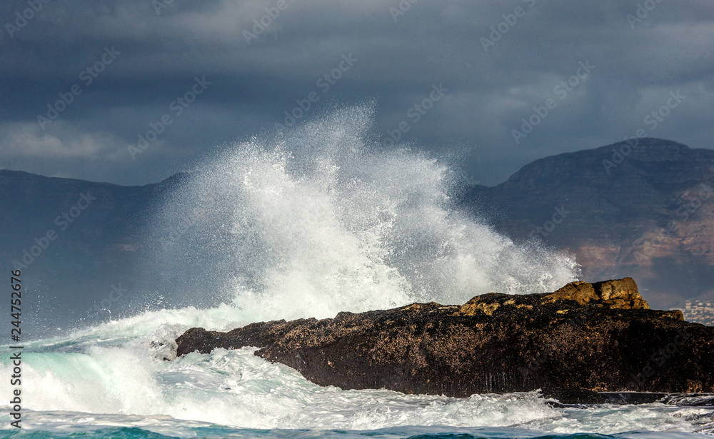 Obraz premium Big waves break on the rocks in the sea against the backdrop of the coastline. Beautiful seascape. A beautiful moment. Very dynamic photo. Cape Town. False Bay. South Africa.