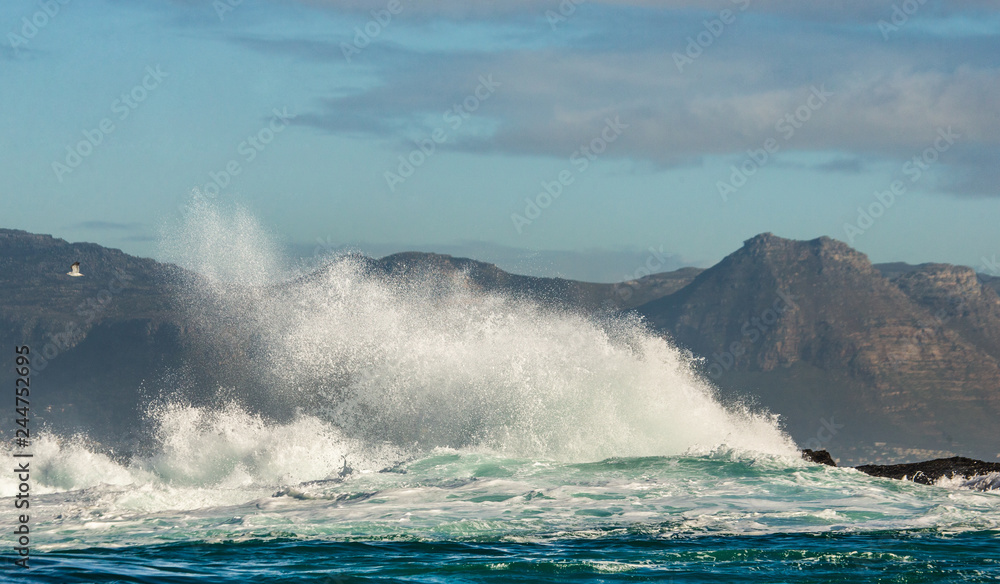 Big waves break on the rocks in the sea against the backdrop of the coastline. Beautiful seascape.  A beautiful moment. Very dynamic photo. Cape Town. False Bay. South Africa.