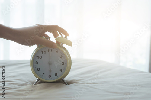 woman hand turn off or snoozing alarm clock waking up on morning in bedroom with sun light.