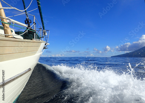 Along the hull toward the bow of a cream and light teak sailboat as it cuts through a dark blue sea creating a smooth wake and white cap curl toward the tip of an island under a blue sky. Copy space.