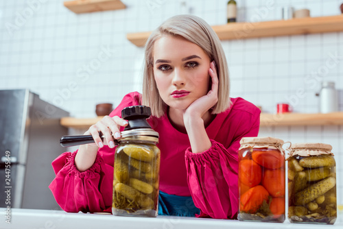 beautiful fashionable housewife looking at camera and holding seamer with jars of pickled vegetables on kitchen counter photo