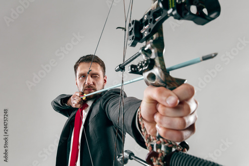 Businessman aiming at target with bow and arrow, isolated on gray studio background. The business, goal, challenge, competition, achievement concept