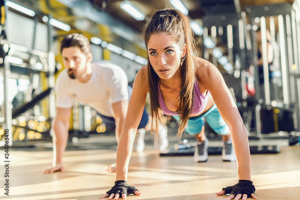 Close up of beautiful Caucasian woman doing push-ups in gym. In background her personal trainer showing exercises.