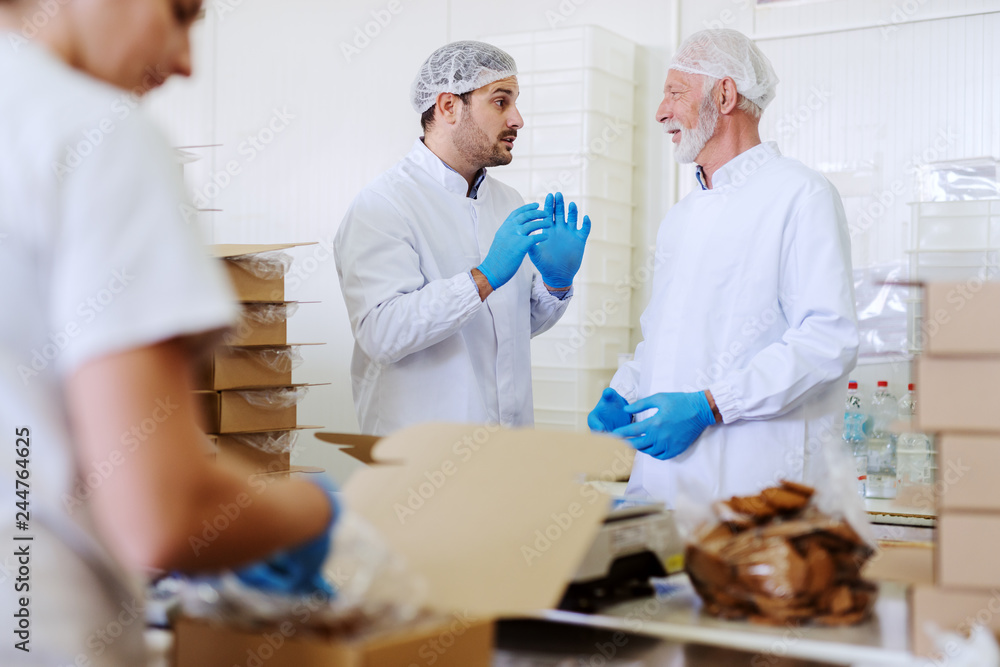 Fototapeta premium Two workers in sterile white uniforms packing cookies and talking while standing in food factory.