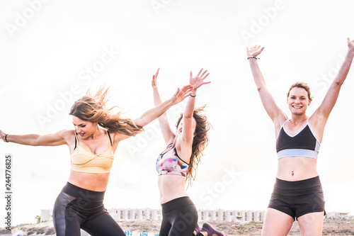 Three young beautiful active athlete girls jumping for success and satisfaction after an outdoor beach workout together in friendship - white clear sky in background