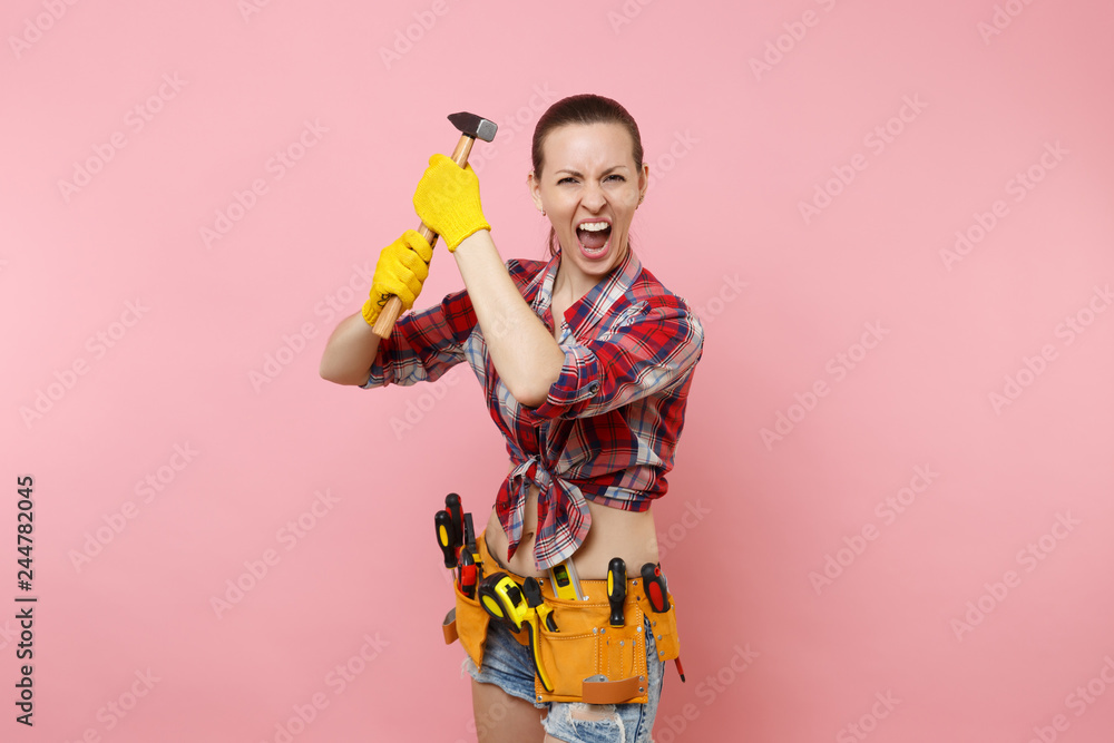 Strong young handyman woman wearing plaid shirt, denim shorts, kit tools belt full of different instruments hammer isolated on pink background. Female in male work. Renovation and occupation concept Strong young handyman woman wearing plaid shirt, denim shorts, kit tools belt full of different instruments hammer isolated on pink background. Female in male work. Renovation and occupation concept