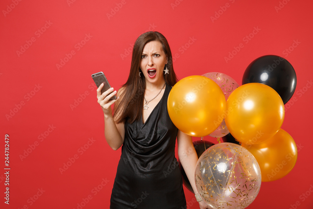 Fototapeta premium Irritated young girl in black dress swearing holding air balloons using mobile phone while celebrating isolated on red background. Valentine's Day Happy New Year birthday mockup holiday party concept.